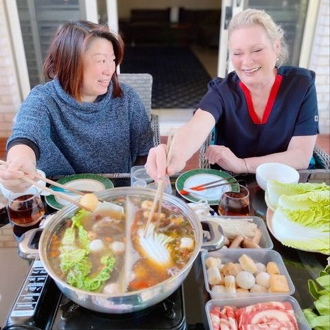 Two friends enjoying hot pot at home, with the Caucasian trying hot pot for the first time. The Asian friend is teaching her how to enjoy hot pot. Both are picking out ingredients from the hot pot broths.