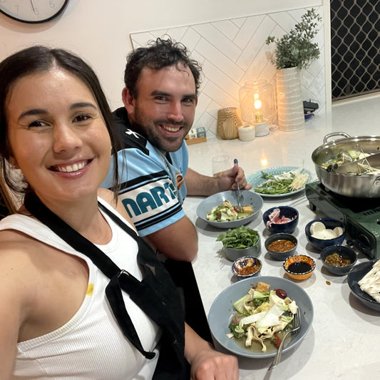 A couple enjoying hot pot at home for date night, looking into the camera. With hot pot ingredients in their bowls.